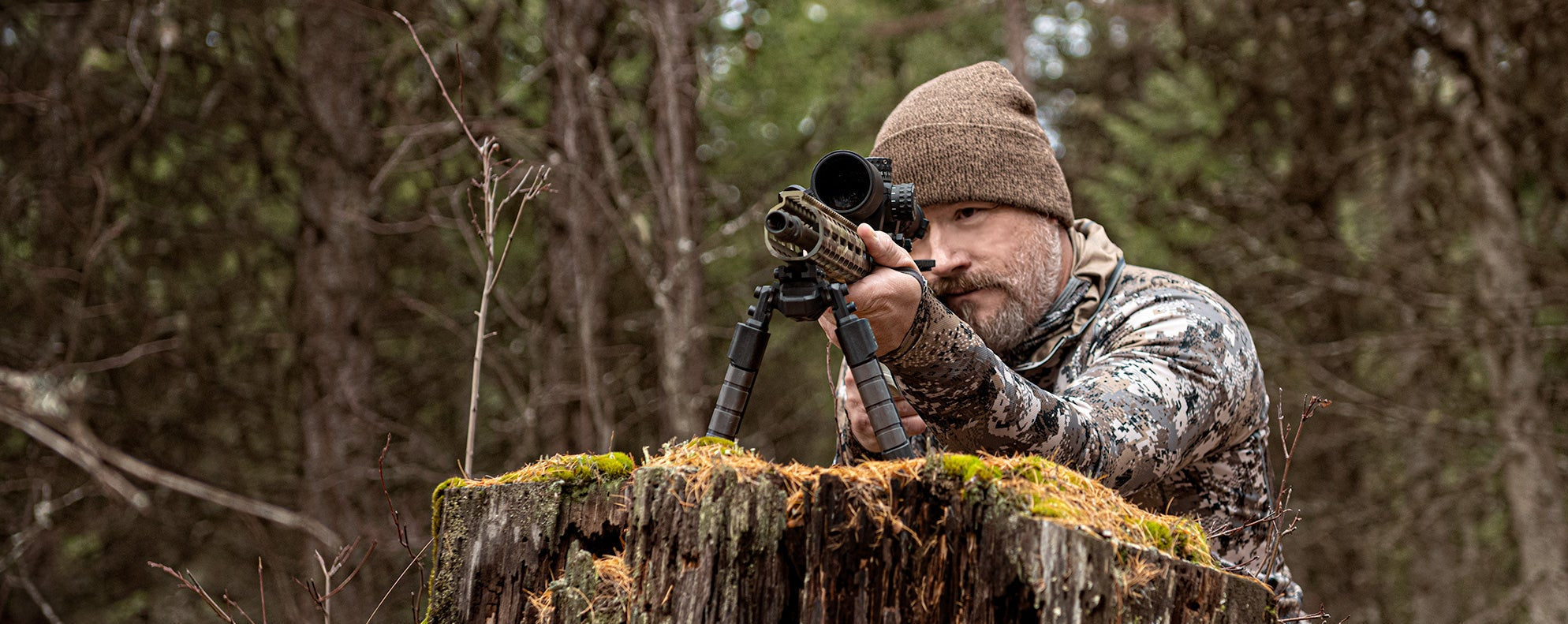 Man in camouflage gear using a rifle with a scope on a tree stump in a forest.